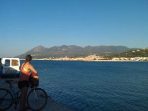 Woman on a bicycle looks across a bay to another Greek island