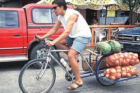 Man carrying 2 watermelons, 2 bags of onions, and a box of potatoes on his bike.